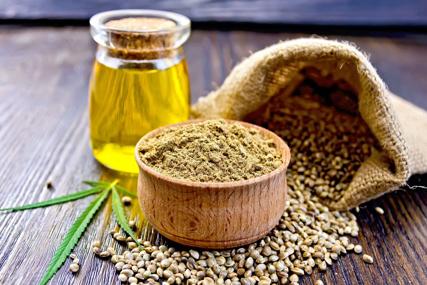 Hemp Flour in a wooden bowl, hemp seed in a bag and on the table, hemp oil in a glass jar, hemp leaf on the background of wooden boards